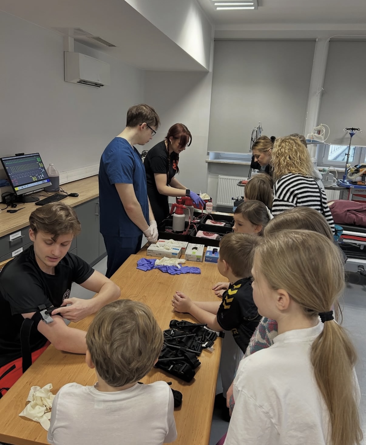 Dzieci uczestniczą w zajęciach warsztatowych, obserwując pokaz związany z pierwszą pomocą lub procedurami medycznymi. Osoby prowadzące prezentują sprzęt i techniki przy stole z materiałami, a jedna z osób demonstruje użycie opaski uciskowej. W tle widoczne są urządzenia medyczne, monitor parametrów oraz wyposażenie sali szkoleniowej.