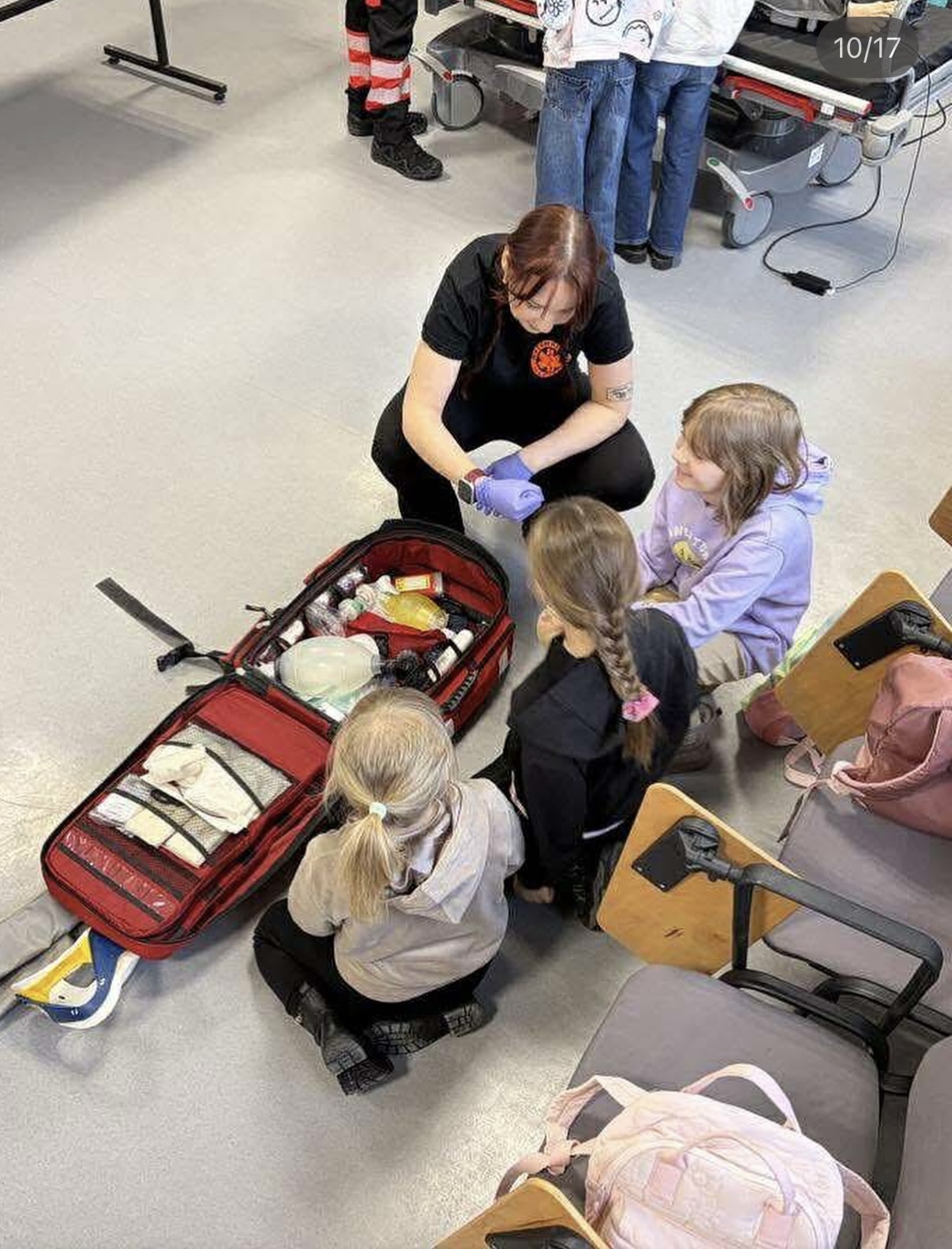 Osoba prowadzi zajęcia edukacyjne z dziećmi, prezentując zawartość zestawu pierwszej pomocy. Dzieci siedzą na podłodze i uważnie obserwują pokaz. W tle widoczne są elementy wyposażenia medycznego, nosze oraz krzesła z plecakami, co wskazuje na salę szkoleniową.