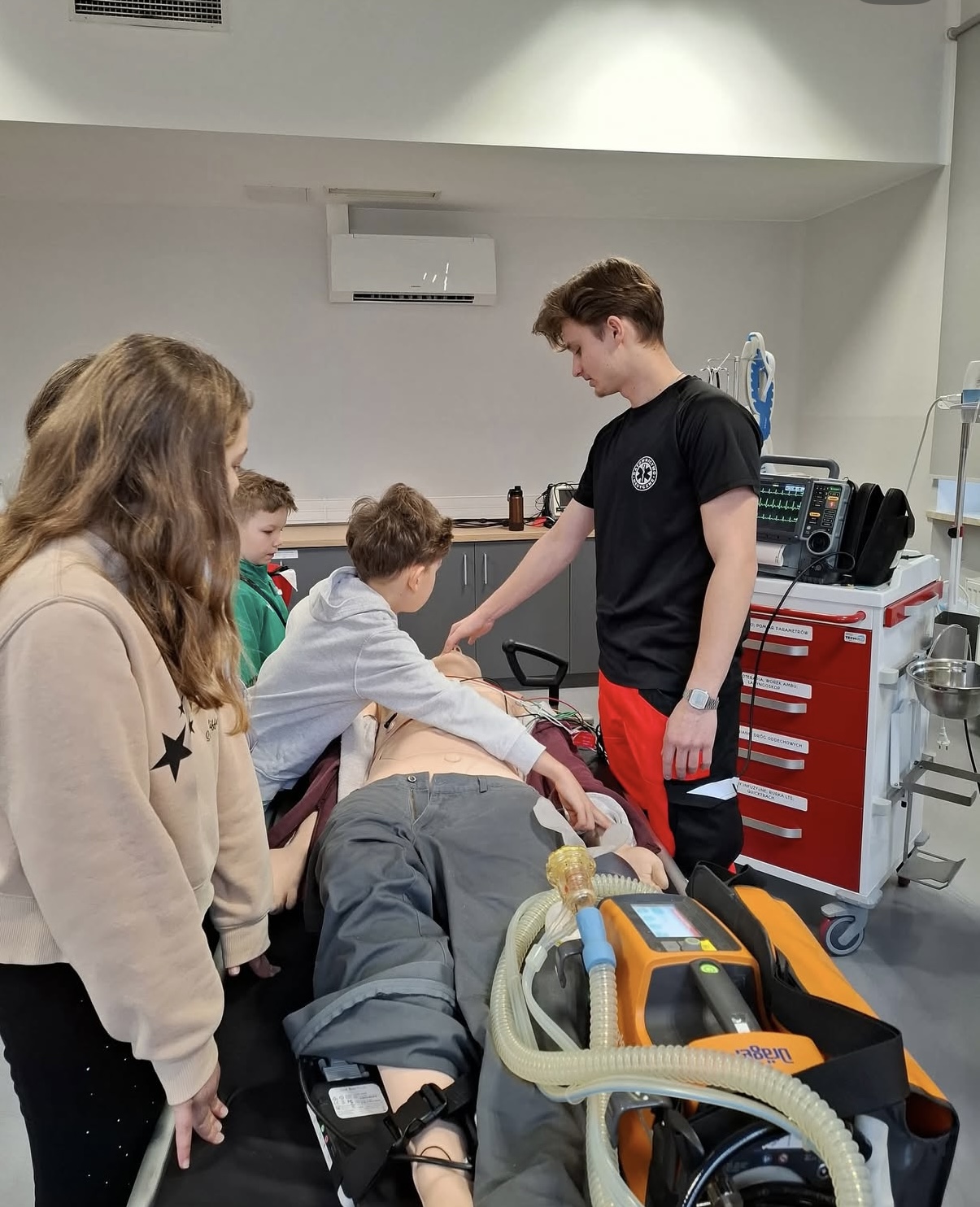 Grupa uczestników bierze udział w zajęciach w sali szkoleniowej o charakterze medycznym, skupiając się wokół fantomu pacjenta na łóżku. Osoba prowadząca demonstruje procedury lub elementy pierwszej pomocy. W tle widoczne są urządzenia medyczne, wózek ratunkowy oraz monitor, co wskazuje na realistyczne warunki nauki.