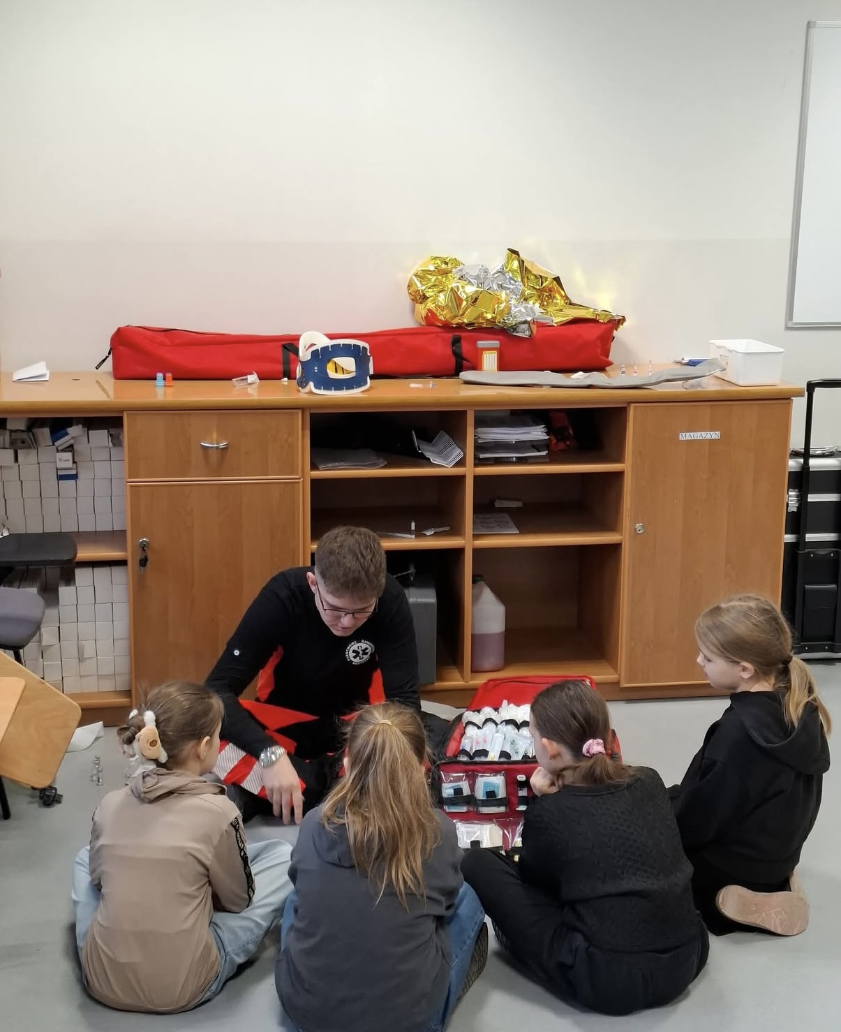 Dzieci siedzą na podłodze w półkolu i uczestniczą w zajęciach edukacyjnych z zakresu pierwszej pomocy. Osoba prowadząca prezentuje zawartość otwartej torby ratunkowej z materiałami medycznymi. W tle widoczna jest szafka z dodatkowymi elementami wyposażenia, co wskazuje na praktyczny charakter szkolenia.