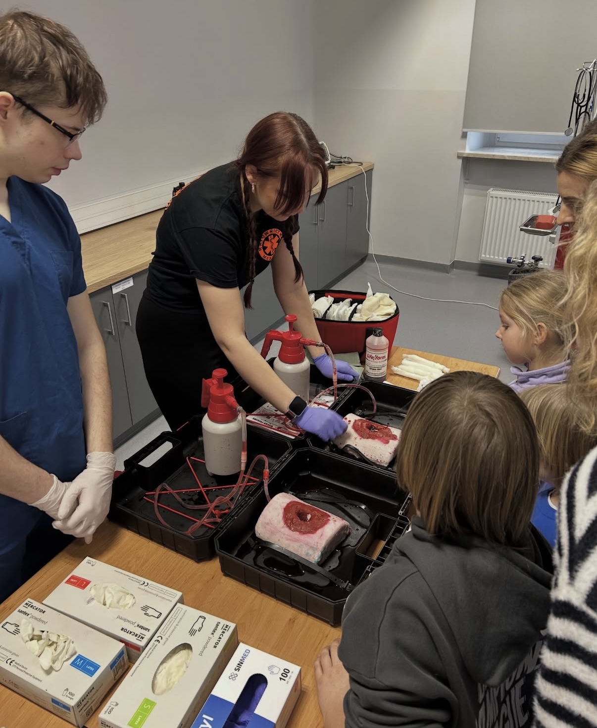 Grupa uczestników stoi przy stole w sali szkoleniowej, obserwując demonstrację procedury medycznej z wykorzystaniem modeli symulujących urazy. Na stole znajdują się zestawy szkoleniowe, rękawice oraz inne materiały medyczne. Elementy z płynem i przewodami odwzorowują krążenie krwi, co podkreśla praktyczny charakter zajęć.