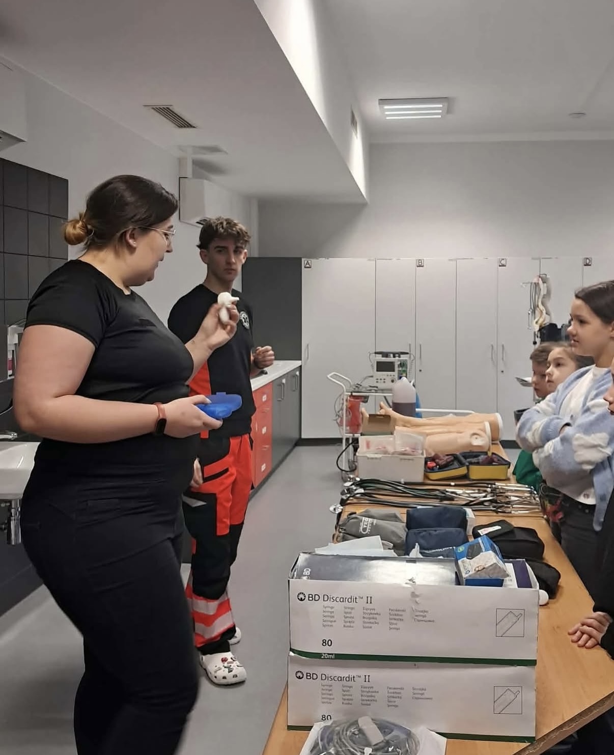 Zajęcia szkoleniowe w sali o charakterze medycznym, podczas których osoba prowadząca prezentuje sprzęt i omawia jego zastosowanie przy stole z materiałami. Uczestnicy, w tym dzieci, obserwują demonstrację. Na stole znajdują się zestawy i narzędzia medyczne, a wnętrze wyposażone jest w nowoczesne szafki i oświetlenie.