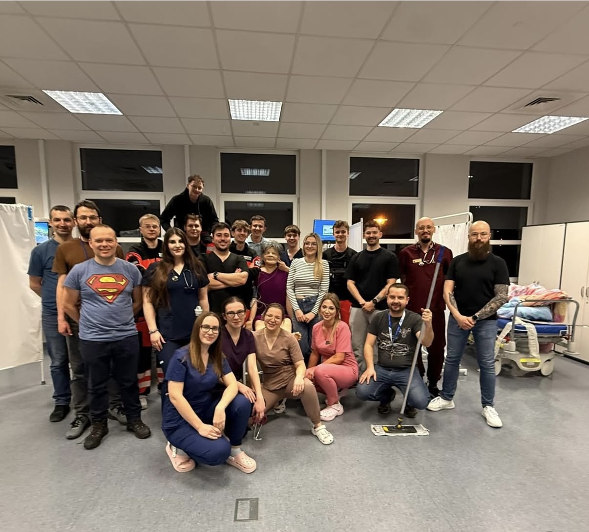 Grupa uczestników zgromadzona jest w pomieszczeniu o charakterze szkoleniowym lub integracyjnym, pozując do wspólnego zdjęcia. Wnętrze jest dobrze oświetlone, z widocznymi oknami, przegrodą oraz elementami wyposażenia przypominającymi salę medyczną lub dydaktyczną.