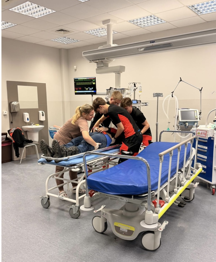 Personel medyczny udziela pomocy pacjentowi leżącemu na łóżku szpitalnym w sali przypominającej oddział ratunkowy. Wokół znajdują się urządzenia medyczne, w tym monitor parametrów oraz stojaki do kroplówek. Scena przedstawia działania zespołu w standardowym środowisku klinicznym.