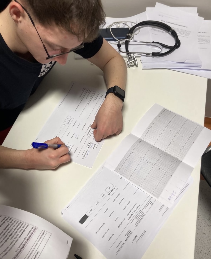 Osoba siedzi przy biurku i sporządza notatki na dokumentach zawierających tabele i wykresy. Na blacie znajdują się rozłożone materiały, stetoskop oraz stos papierów, co wskazuje na pracę o charakterze medycznym lub akademickim.