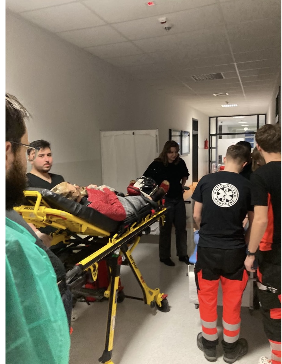 Zespół medyczny udziela pomocy pacjentowi leżącemu na noszach w korytarzu szpitalnym. Wokół znajdują się osoby zaangażowane w działania ratunkowe, a przestrzeń oświetlona jest światłem sufitowym. W tle widoczne są elementy infrastruktury szpitalnej, takie jak drzwi i kurtyna oddzielająca stanowiska.