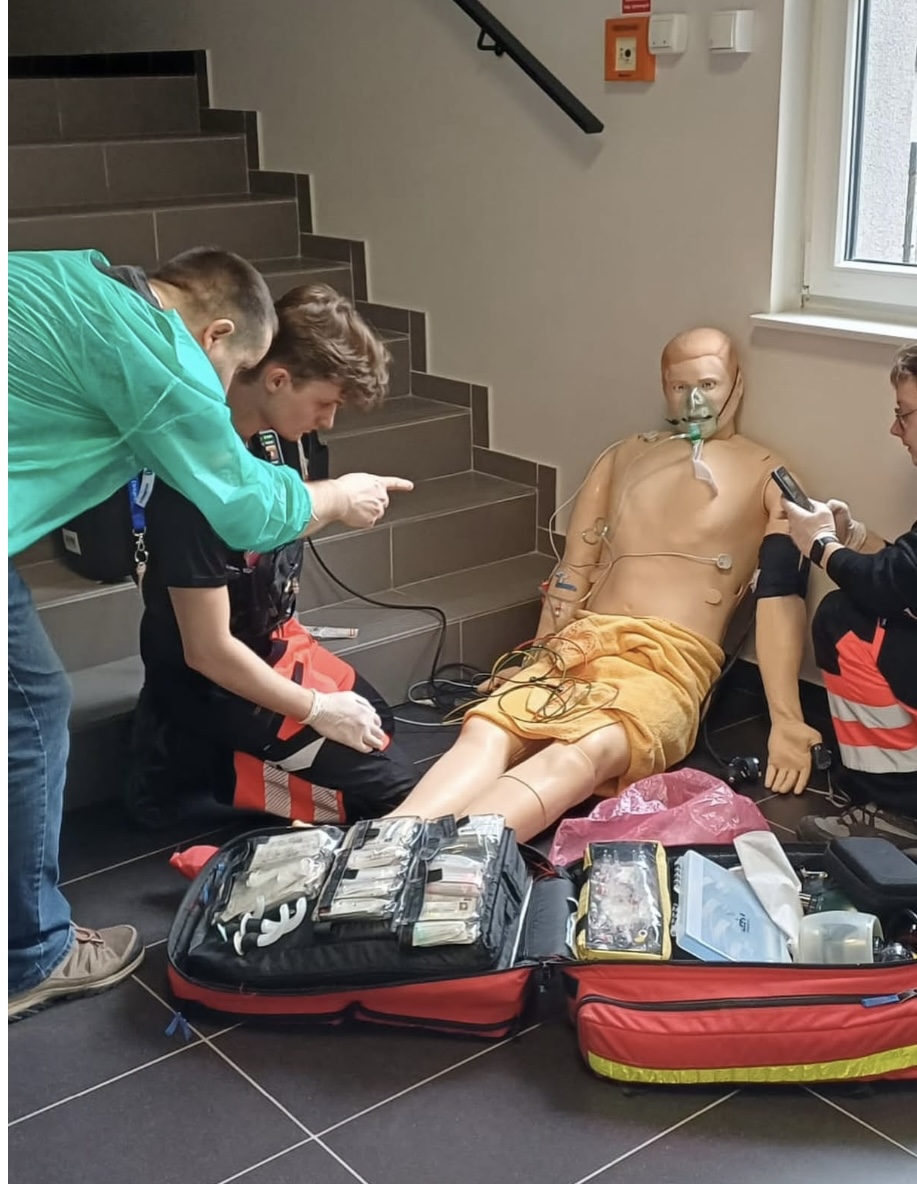 Uczestnicy biorą udział w szkoleniu medycznym, udzielając pomocy fantomowi pacjenta siedzącemu przy schodach. Osoba prowadząca wskazuje kolejne czynności, a pozostali wykonują działania z użyciem sprzętu z otwartych zestawów ratunkowych. Fantom wyposażony jest w maskę tlenową i czujniki, co umożliwia realistyczną symulację.