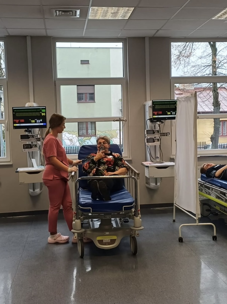 Sala szpitalna z dwoma łóżkami wyposażonymi w monitory medyczne. Jedna z osób udziela pomocy pacjentowi siedzącemu na łóżku, który korzysta z maski oddechowej. W tle widoczne są duże okna z widokiem na zewnątrz oraz zasłona oddzielająca stanowiska, co wskazuje na standardowe środowisko kliniczne.