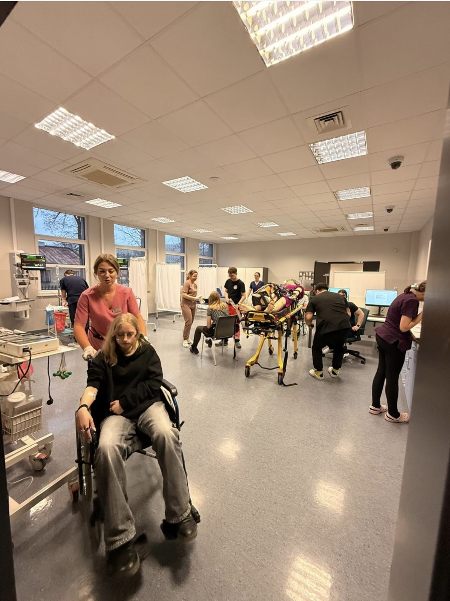 Sala szpitalna, w której personel medyczny prowadzi różne działania opiekuńcze. Na pierwszym planie transportowany jest pacjent na wózku, a w tle widoczne są stanowiska pracy, łóżko szpitalne oraz osoby wykonujące czynności przy komputerze. Pomieszczenie jest dobrze oświetlone, z dużymi oknami i zasłonami oddzielającymi przestrzeń.