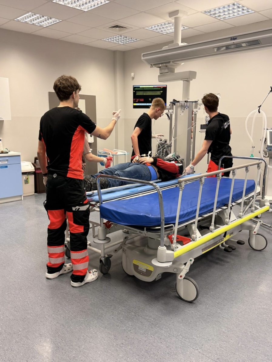 Uczestnicy biorą udział w symulacji medycznej w sali przypominającej oddział szpitalny, pracując przy łóżku z fantomem podłączonym do urządzeń monitorujących. Obok znajduje się monitor parametrów życiowych, a całość wskazuje na ćwiczenia praktyczne w realistycznych warunkach klinicznych.