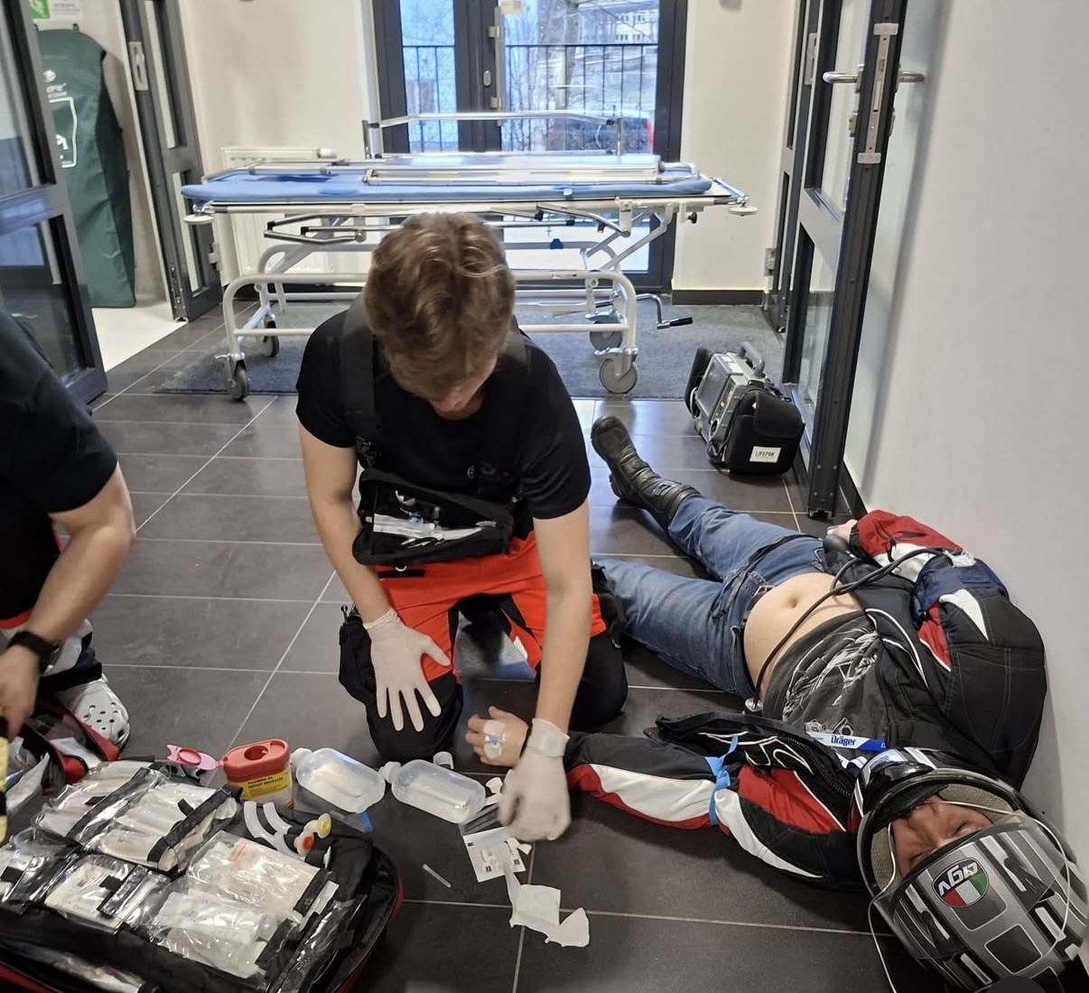 Osoba udziela pomocy poszkodowanemu leżącemu na podłodze, wykonując czynności ratunkowe z użyciem sprzętu z otwartego zestawu medycznego. W pobliżu znajdują się nosze oraz dodatkowe wyposażenie, a cała scena rozgrywa się w przestrzeni przypominającej korytarz budynku, z dostępem do światła dziennego.