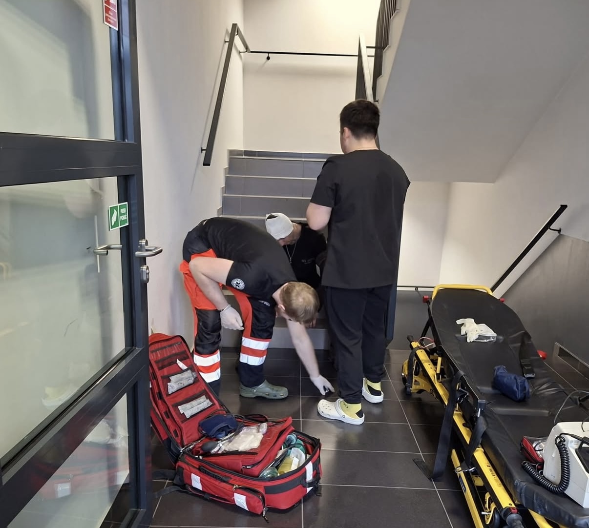 Uczestnicy udzielają pomocy w sytuacji ratunkowej na klatce schodowej, wykonując czynności przy poszkodowanym. Obok znajduje się otwarty zestaw medyczny oraz nosze z przygotowanym sprzętem, co wskazuje na działania interwencyjne.
