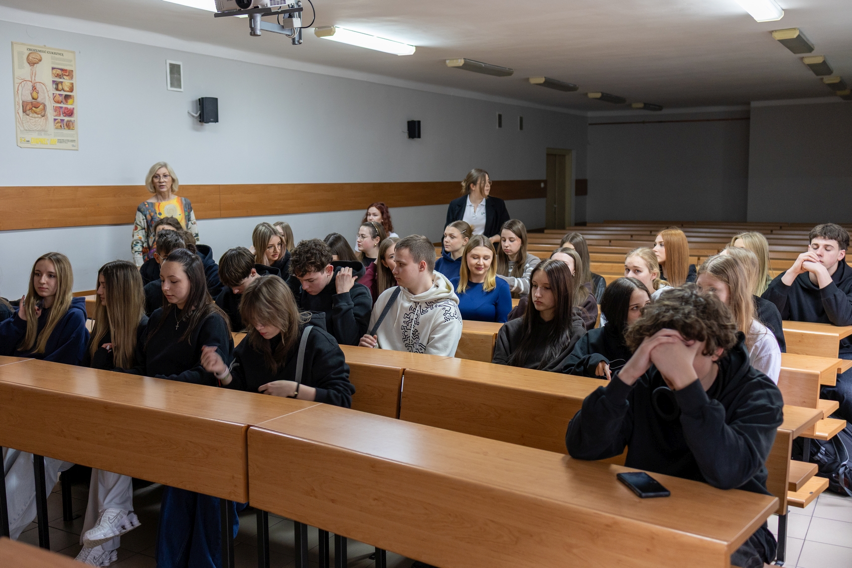 Sala dydaktyczna z rzędami drewnianych ławek, w której uczestnicy biorą udział w zajęciach. Część osób notuje lub słucha, a z tyłu sali stoją osoby prowadzące lub nadzorujące. Na ścianie widoczny jest plakat z ilustracjami anatomicznymi, a nad salą zamontowany jest projektor.