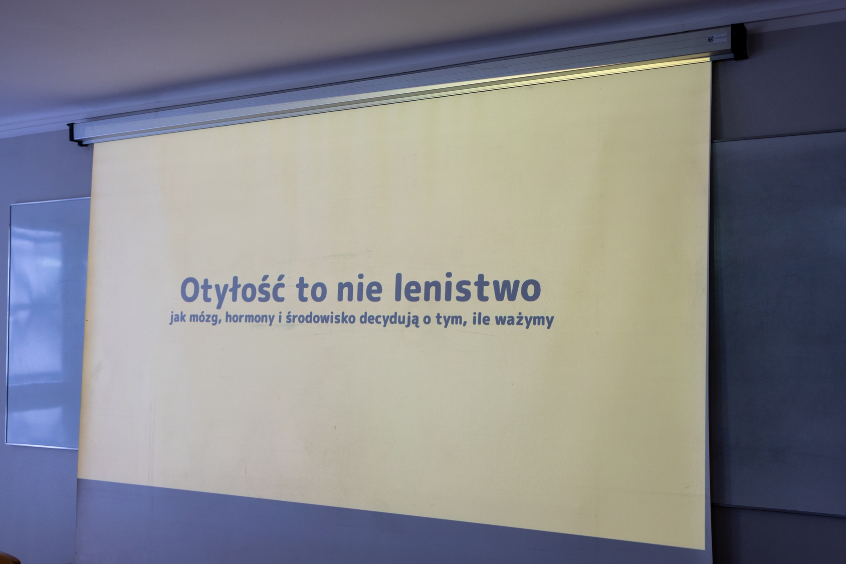 Ekran projekcyjny wyświetla komunikat w języku polskim na jasnym tle. Znajduje się on na ścianie w sali dydaktycznej, obok widoczna jest część tablicy, co wskazuje na środowisko edukacyjne.