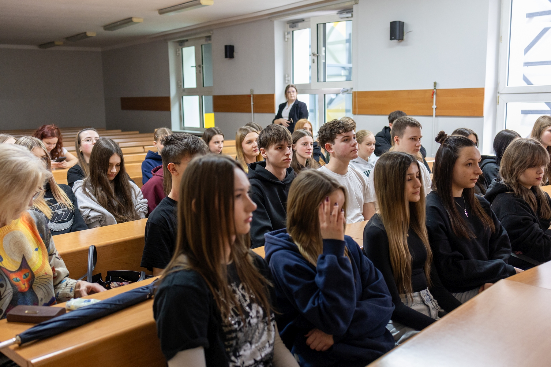 Uczestnicy siedzą w drewnianych ławkach w sali lekcyjnej i obserwują zajęcia prowadzone z przodu. Pomieszczenie jest jasne, oświetlone światłem sufitowym oraz dziennym wpadającym przez duże okna.