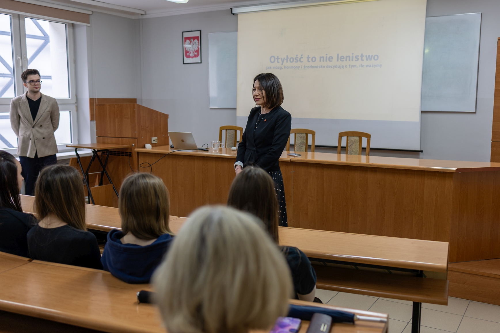 Sala dydaktyczna z drewnianymi ławkami ustawionymi w rzędach, w której uczestnicy siedzą i słuchają wystąpienia prowadzonego z przodu. Przy biurku znajduje się laptop i materiały, a w tle widoczny jest ekran z wyświetlaną prezentacją. Pomieszczenie ma jasne ściany, duże okno oraz elementy wyposażenia, takie jak obraz na ścianie.