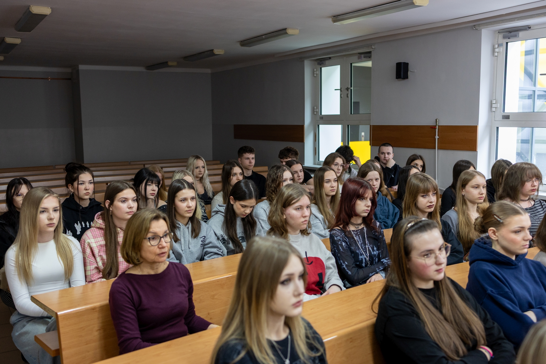 Sala dydaktyczna z rzędami drewnianych ławek, w której uczestnicy siedzą i uczestniczą w zajęciach. Pomieszczenie ma jasne ściany, oświetlenie sufitowe oraz duże okna wpuszczające światło dzienne. Publiczność jest skupiona na przebiegu lekcji, siedząc blisko siebie w kolejnych rzędach.