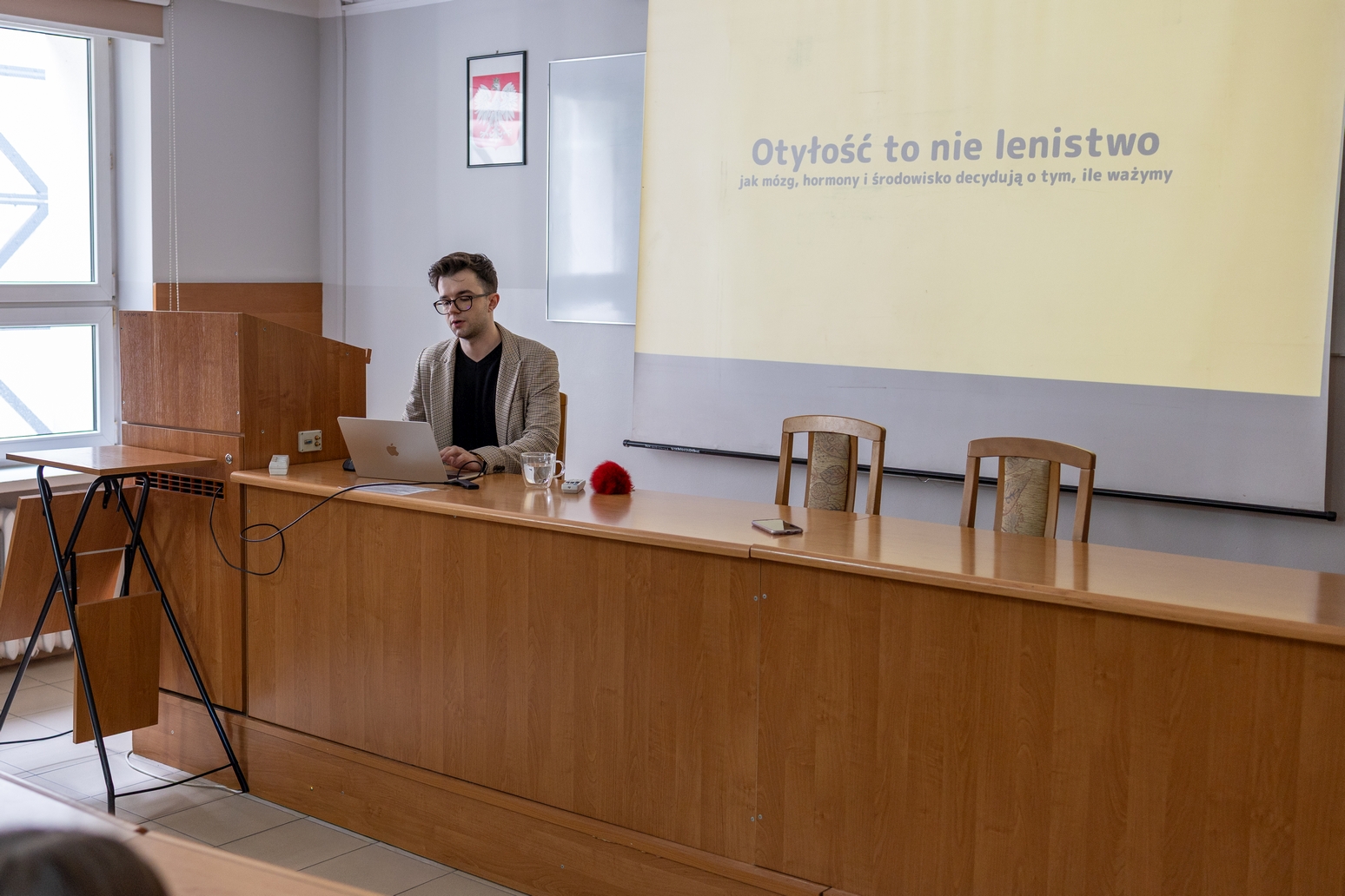 Sala dydaktyczna z biurkiem ustawionym przed ekranem projekcyjnym, na którym wyświetlany jest slajd z tekstem. Przy biurku znajduje się laptop, a za nim ustawione są krzesła. Pomieszczenie ma jasne ściany, okno oraz podstawowe wyposażenie, co wskazuje na środowisko wykładowe lub szkoleniowe.