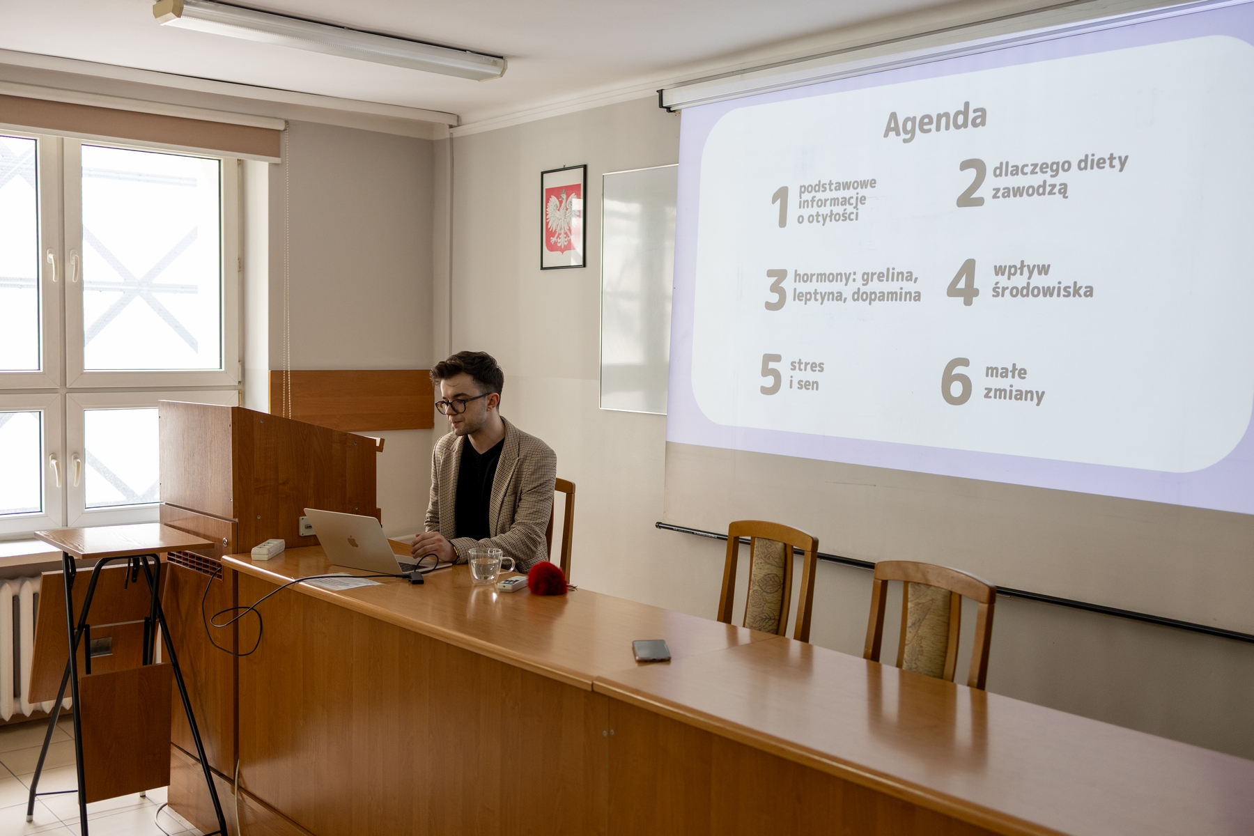 Osoba siedzi przy mównicy i pracuje na laptopie podczas zajęć lub prezentacji. Za nią widoczny jest ekran z wyświetlonym planem lub slajdem. Sala ma jasne ściany, duże okna wpuszczające światło dzienne oraz podstawowe wyposażenie, takie jak stół z materiałami.