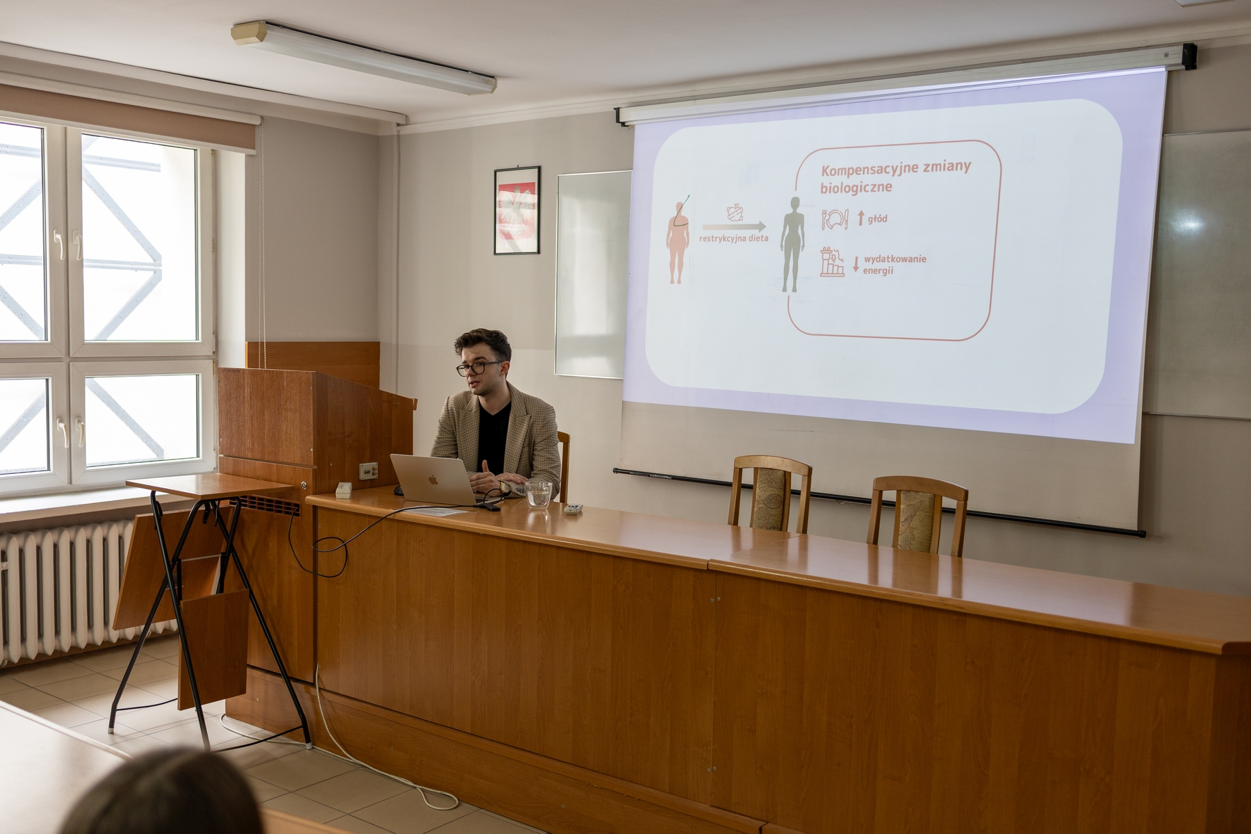 Sala dydaktyczna z biurkiem i mównicą ustawionymi z przodu, gdzie prowadzona jest prezentacja z wykorzystaniem laptopa. Obok znajduje się ekran z wyświetlonym slajdem zawierającym grafikę i tekst w języku polskim. Pomieszczenie jest dobrze oświetlone światłem dziennym z dużego okna, a wnętrze ma jasne ściany i podstawowe wyposażenie.