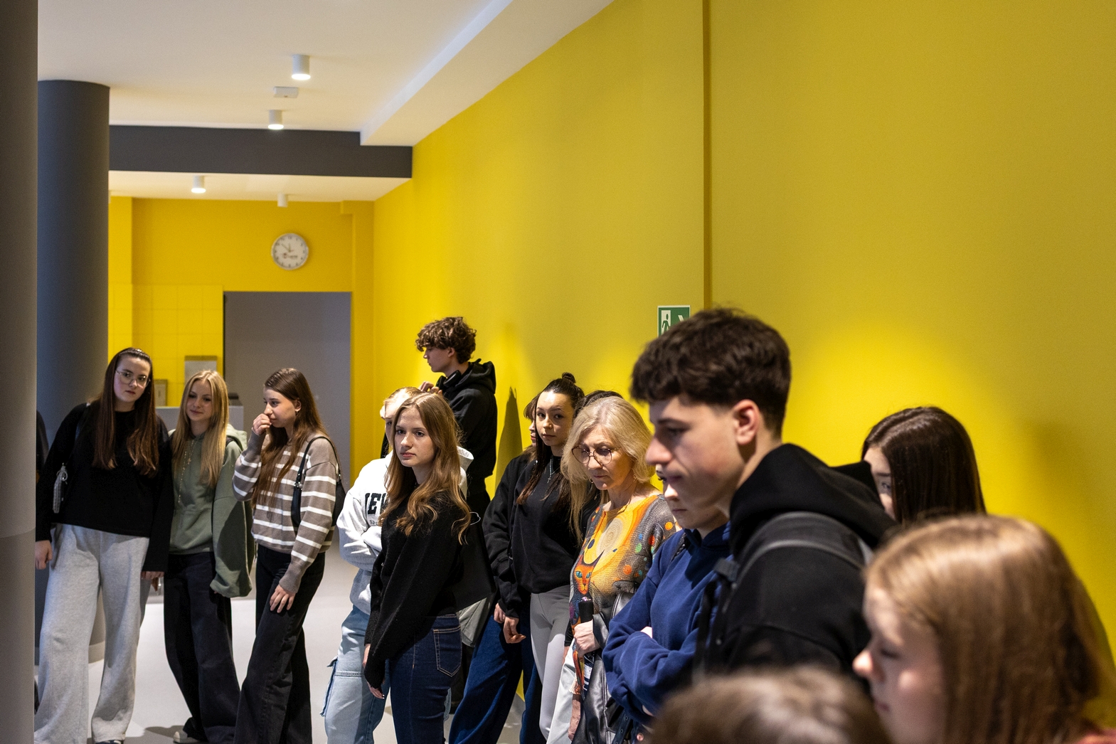 Grupa uczestników stoi w pomieszczeniu przy jasnej ścianie, słuchając lub obserwując coś poza kadrem. Wnętrze ma nowoczesny, minimalistyczny charakter z elementami takimi jak kolumna, zegar oraz oświetlenie sufitowe. Atmosfera jest swobodna, a przestrzeń przypomina miejsce spotkań lub zajęć.