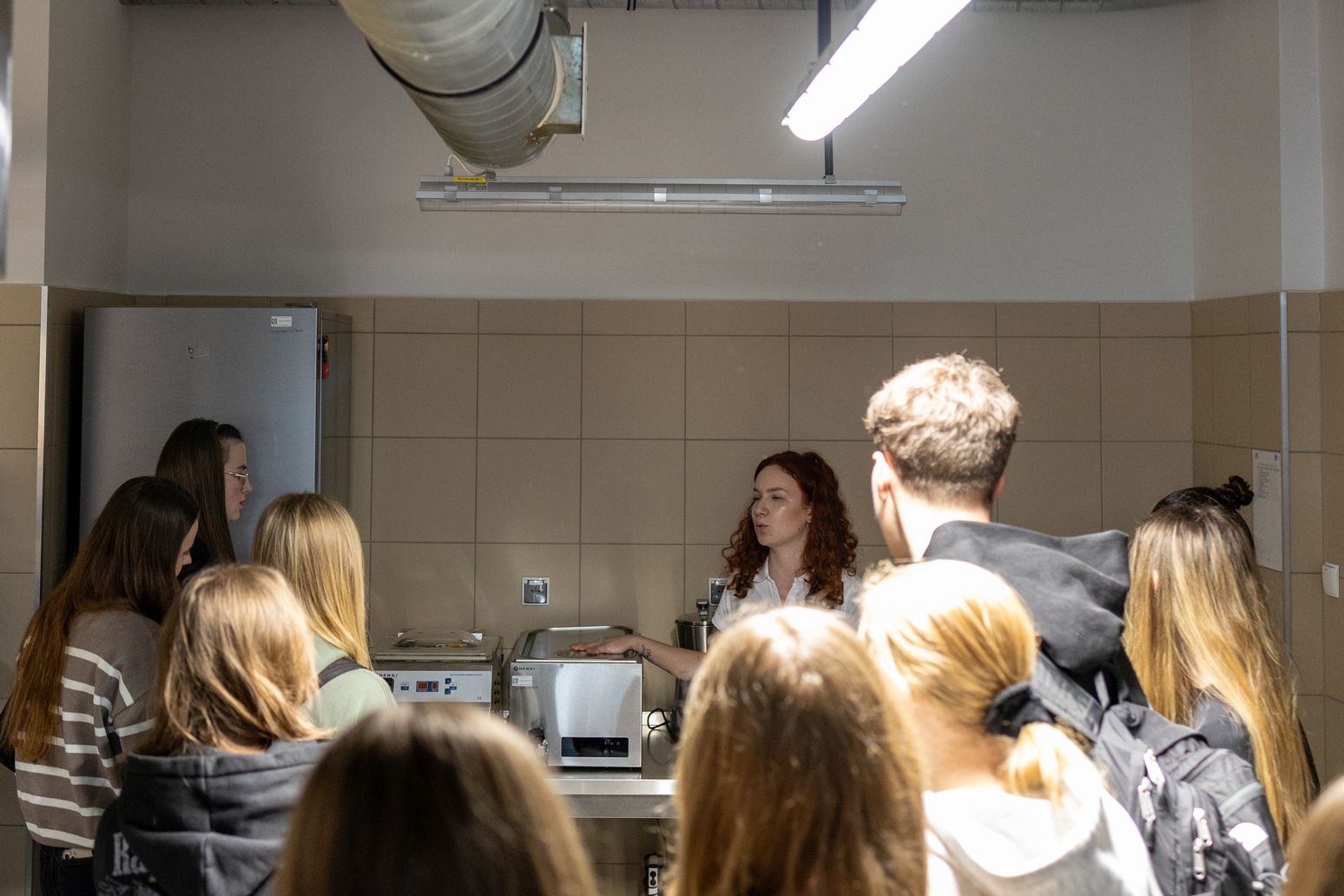Grupa uczestników zgromadzona jest w pomieszczeniu przypominającym zaplecze kuchenne, gdzie jedna z osób prowadzi prezentację lub pokaz przy stanowisku roboczym. Na blacie widoczne są urządzenia kuchenne, a uczestnicy stoją i słuchają, co wskazuje na warsztatowy lub szkoleniowy charakter spotkania.