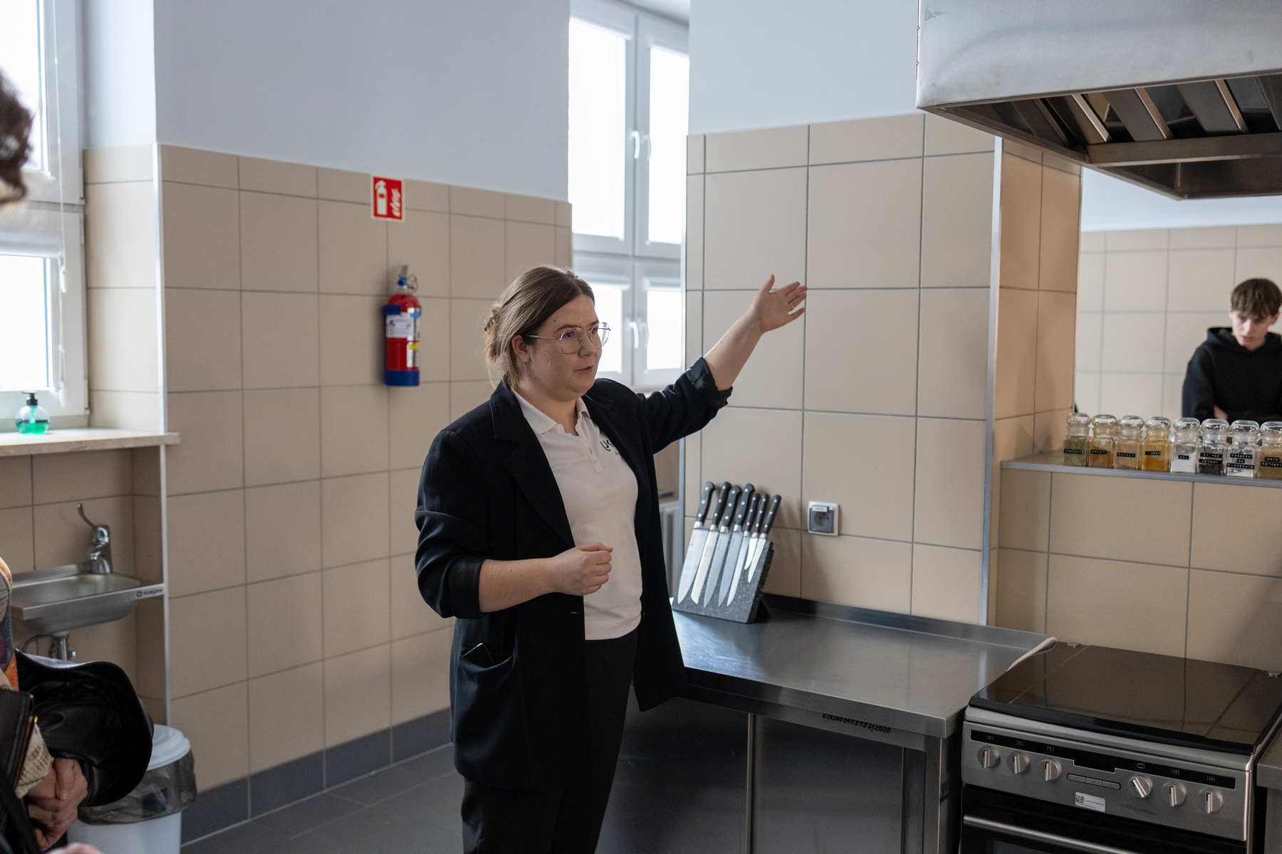 Osoba prowadzi prezentację w profesjonalnej kuchni, stojąc przy stanowisku roboczym. Wnętrze wyposażone jest w blat ze sprzętem kuchennym, przyprawy oraz kuchenkę z okapem. Pomieszczenie ma jasne ściany z kafelków, duże okna oraz elementy wyposażenia, takie jak gaśnica i zlew, co wskazuje na środowisko warsztatowe lub szkoleniowe.