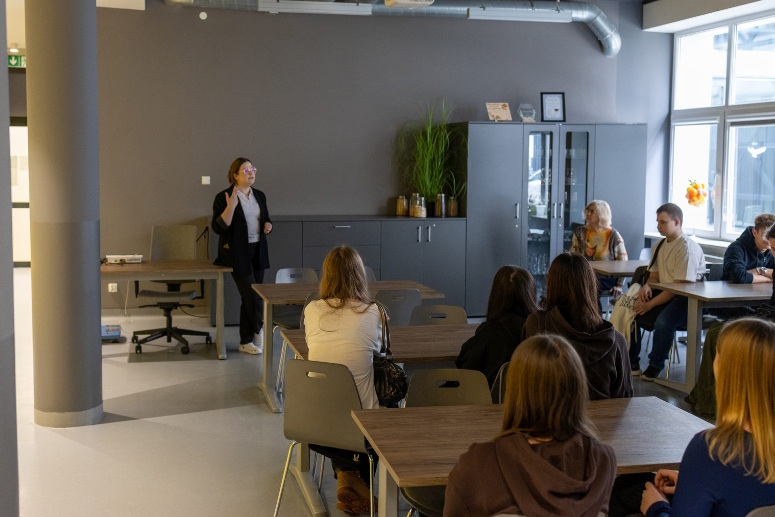 Uczestnicy siedzą przy stołach w sali szkoleniowej i słuchają wystąpienia prowadzonego z przodu. Osoba prowadząca prezentację omawia temat, stojąc przed grupą. Wnętrze ma nowoczesny charakter z jasnymi ścianami, dużymi oknami wpuszczającymi światło dzienne oraz szafą z przeszklonymi drzwiami i roślinami.