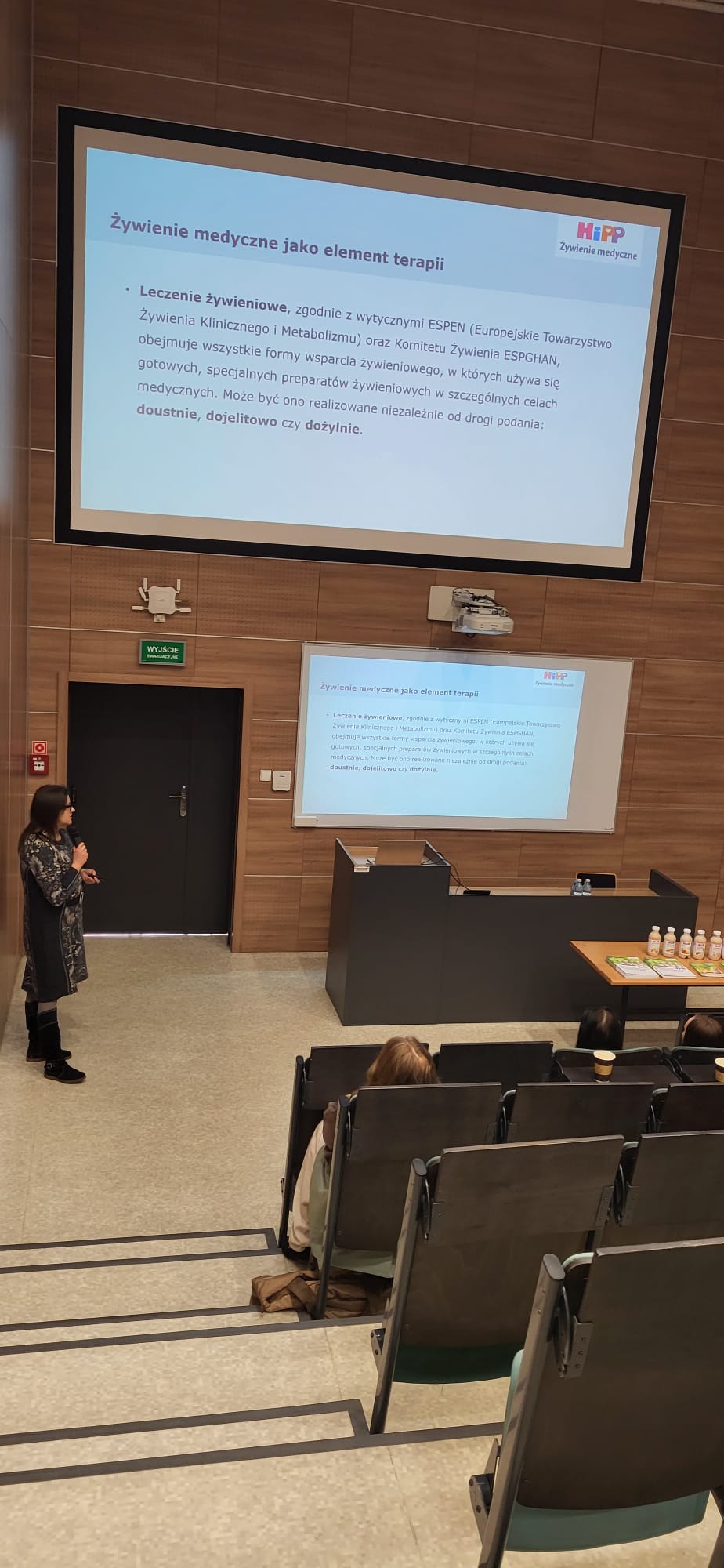 Sala wykładowa z rzędami stopniowanych siedzeń skierowanych w stronę sceny. Z przodu osoba prowadzi wystąpienie przy mównicy z mikrofonem. Za nią znajdują się dwa ekrany z wyświetlonym slajdem prezentacji zawierającym tytuł i punkty tekstowe. Obok mównicy stoi stół z butelkami i materiałami informacyjnymi, a wnętrze sali wykończone jest drewnianymi panelami.