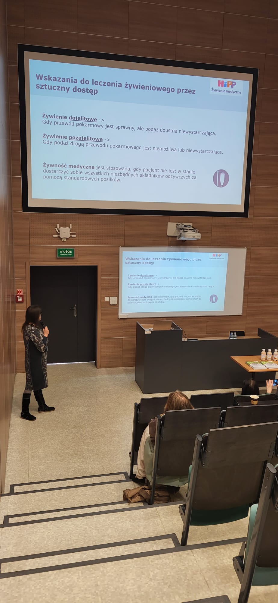 Sala wykładowa z rzędami stopniowanych siedzeń oraz drewnianymi panelami na ścianach. Z przodu widoczne są dwa ekrany z wyświetlonym slajdem prezentacji w języku polskim dotyczącym terapii żywieniowej. Jedna z osób prowadzi wystąpienie z mikrofonem, stojąc przy ekranie, a obok znajduje się stół z materiałami oraz produktami prezentowanymi podczas spotkania. W tle widoczne są drzwi ewakuacyjne i projektor zamontowany nad ekranem.