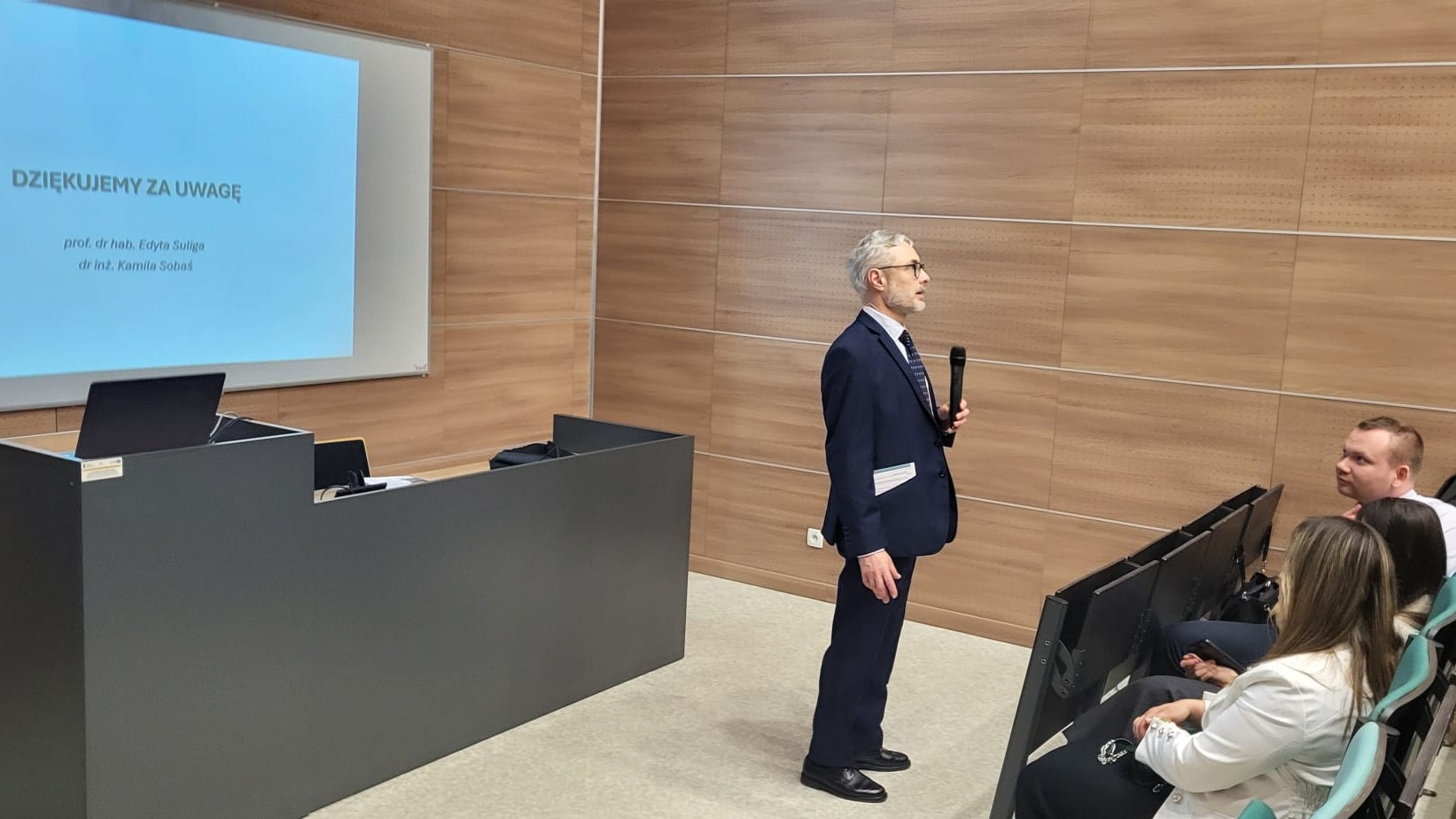 Sala wykładowa o nowoczesnym, akademickim charakterze z drewnianymi panelami na ścianach. Z przodu znajduje się mównica z laptopem oraz ekran z wyświetloną prezentacją. Osoba prowadzi wystąpienie z mikrofonem, a uczestnicy siedzą w rzędach i słuchają wykładu.