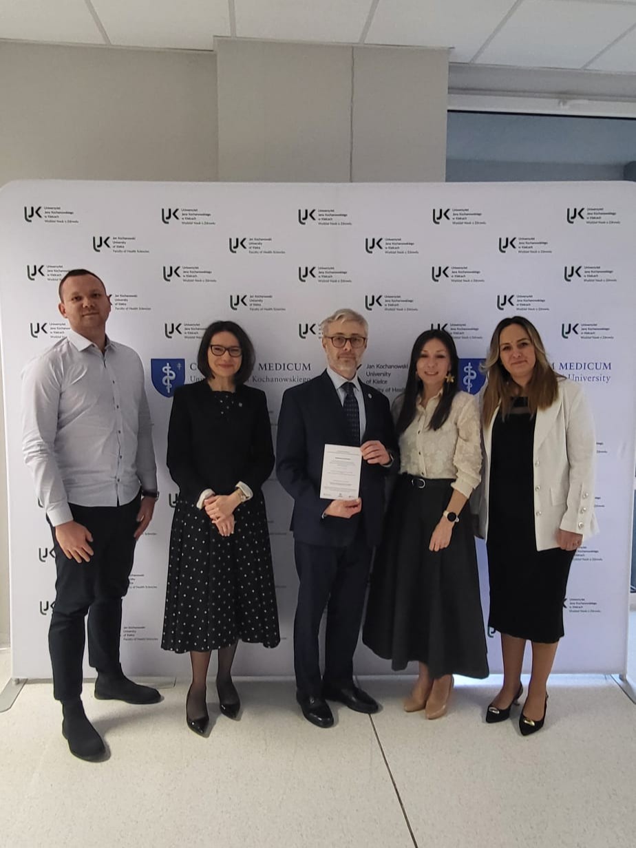 Grupa uczestników stoi przed ścianką z logotypami uczelni, pozując do zdjęcia podczas wydarzenia. Jedna z osób trzyma certyfikat lub dyplom. Wnętrze ma jasną podłogę i sufit z panelami, co wskazuje na oficjalny charakter spotkania.