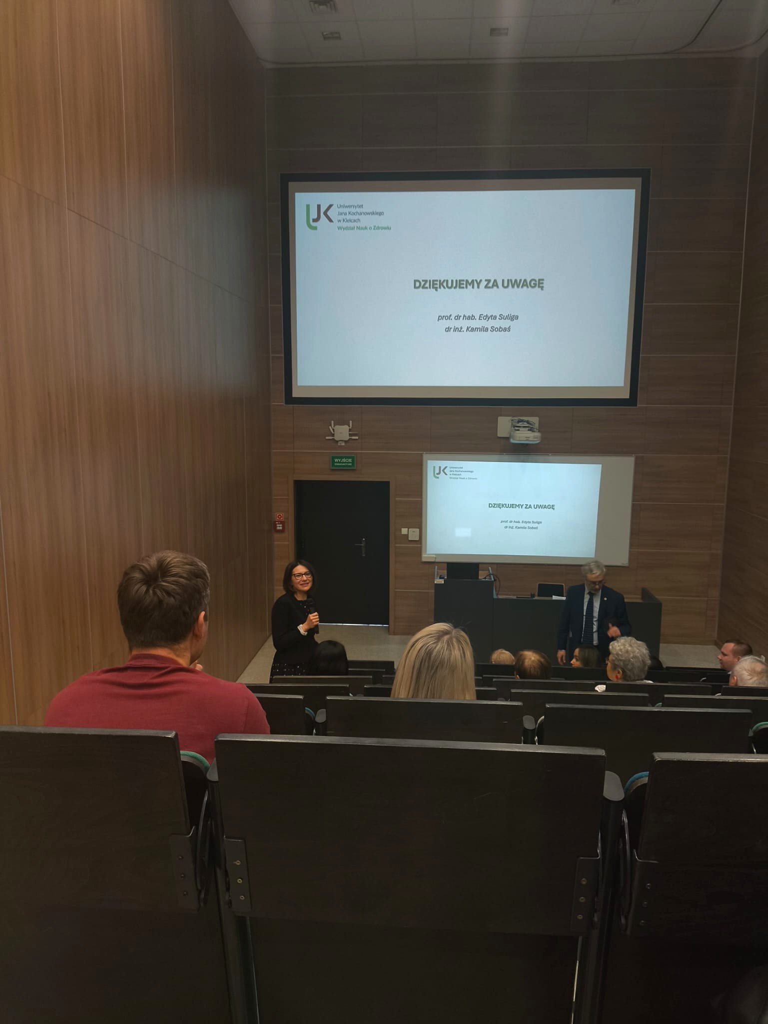 Sala wykładowa z drewnianymi panelami, widziana od tyłu w kierunku sceny. Uczestnicy siedzą w rzędach, a z przodu widoczne są dwa ekrany z wyświetlaną prezentacją. Osoba prowadzi wystąpienie z mikrofonem, a obok znajduje się druga osoba angażująca publiczność. W tle widoczne są drzwi oraz elementy wyposażenia sali.