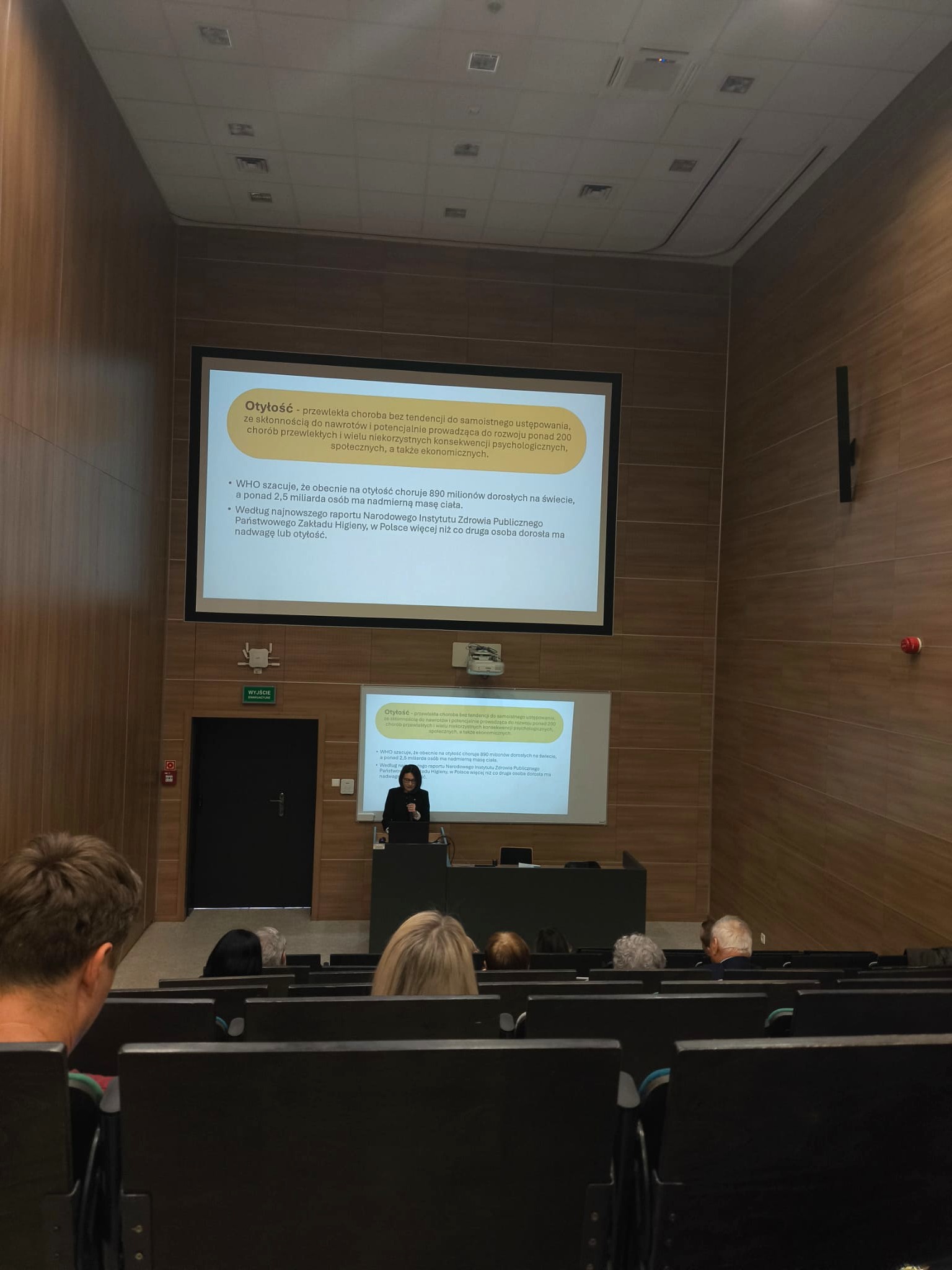 Sala wykładowa z drewnianymi panelami na ścianach i rzędami siedzeń skierowanych w stronę sceny. Z przodu znajduje się ekran z wyświetloną prezentacją, a przy mównicy prowadzony jest wykład. Pomieszczenie ma oświetlenie sufitowe oraz elementy wentylacyjne, co podkreśla jego akademicki charakter.