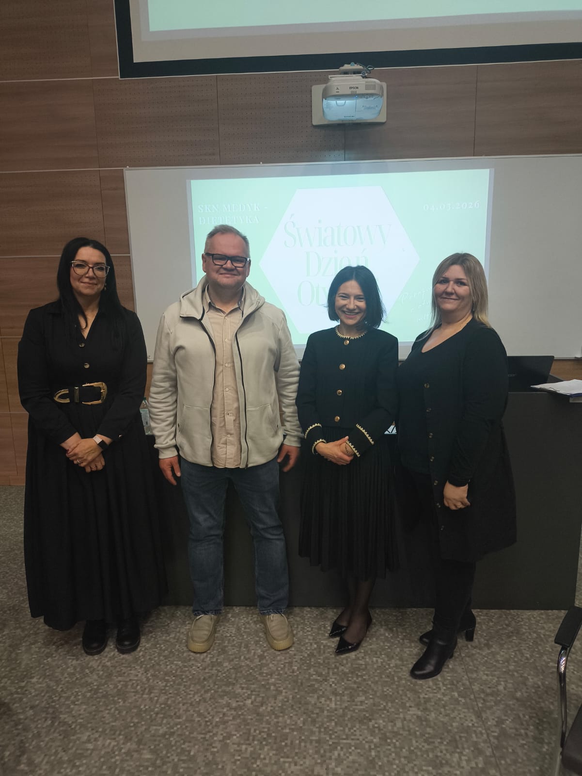 Obraz przedstawia osoby stojące przed białym ekranem projekcyjnym z wyświetlonym na nim sześciokątem zawierającym tekst. Pomieszczenie ma drewniane ściany, sufitowy projektor nad ekranem oraz podłogę z szarym wzorzystym dywanem.