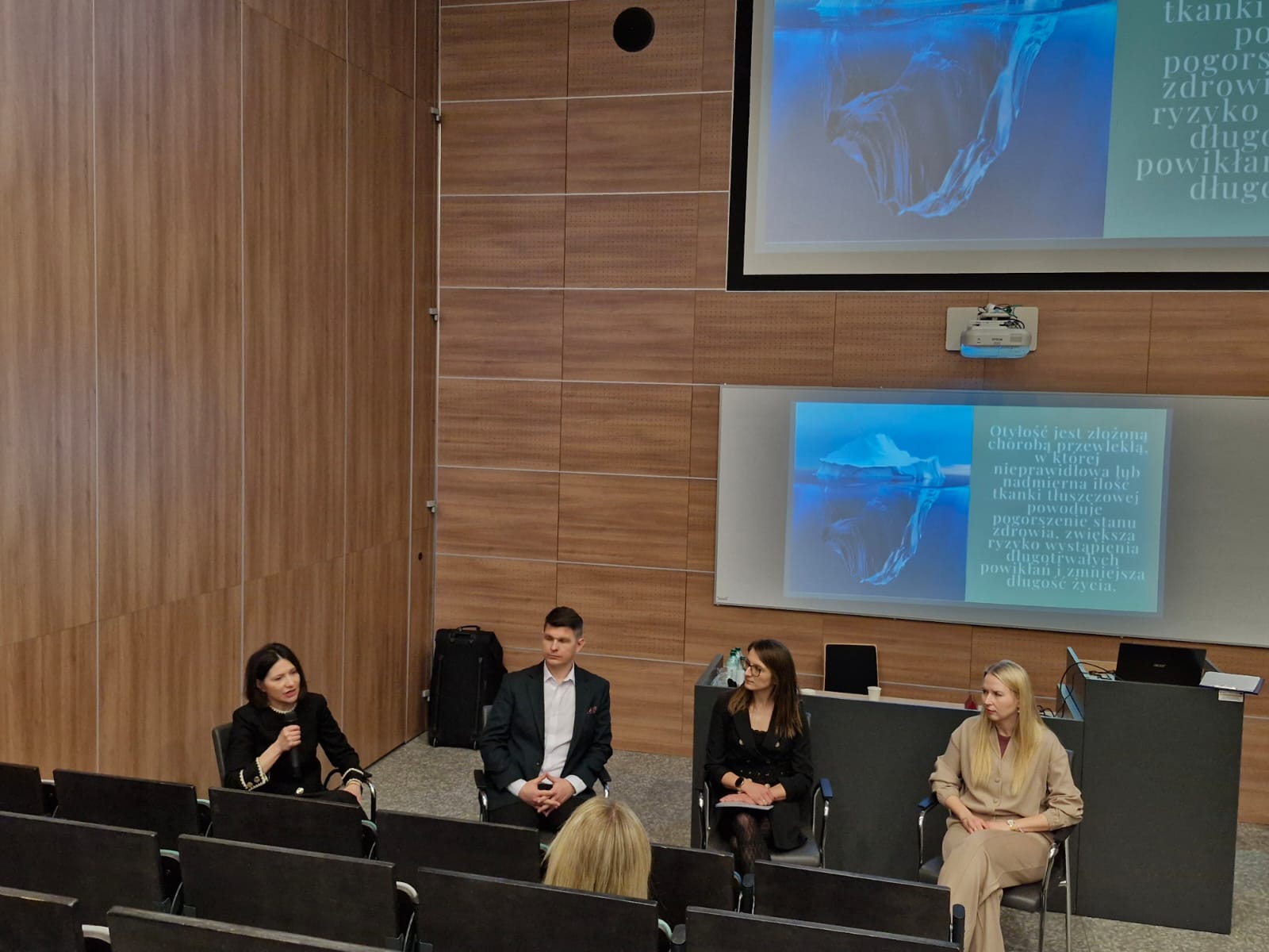 Obraz przedstawia salę wykładową z drewnianymi ścianami i szarym dywanem. Z przodu siedzą osoby zwrócone w stronę publiczności, prowadząc dyskusję. Nad nimi projektor wyświetla na ekranie obraz góry lodowej z nałożonym tekstem. W sali znajdują się czarne krzesła ustawione dla widowni.