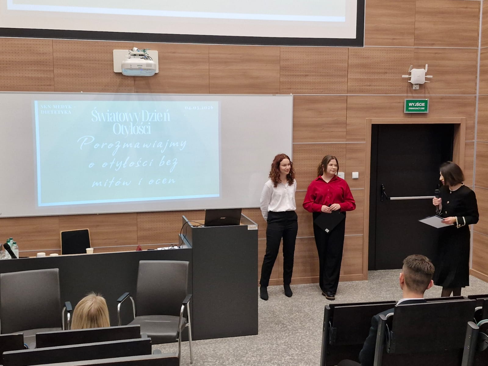 Obraz przedstawia salę wykładową z drewnianymi ścianami. Z przodu, przy mównicy i ekranie projekcyjnym z prezentacją, trwa rozmowa lub wystąpienie z użyciem mikrofonu. W sali znajdują się krzesła dla uczestników oraz biurko z różnymi przedmiotami. Całość sugeruje formalne spotkanie, takie jak konferencja lub seminarium.