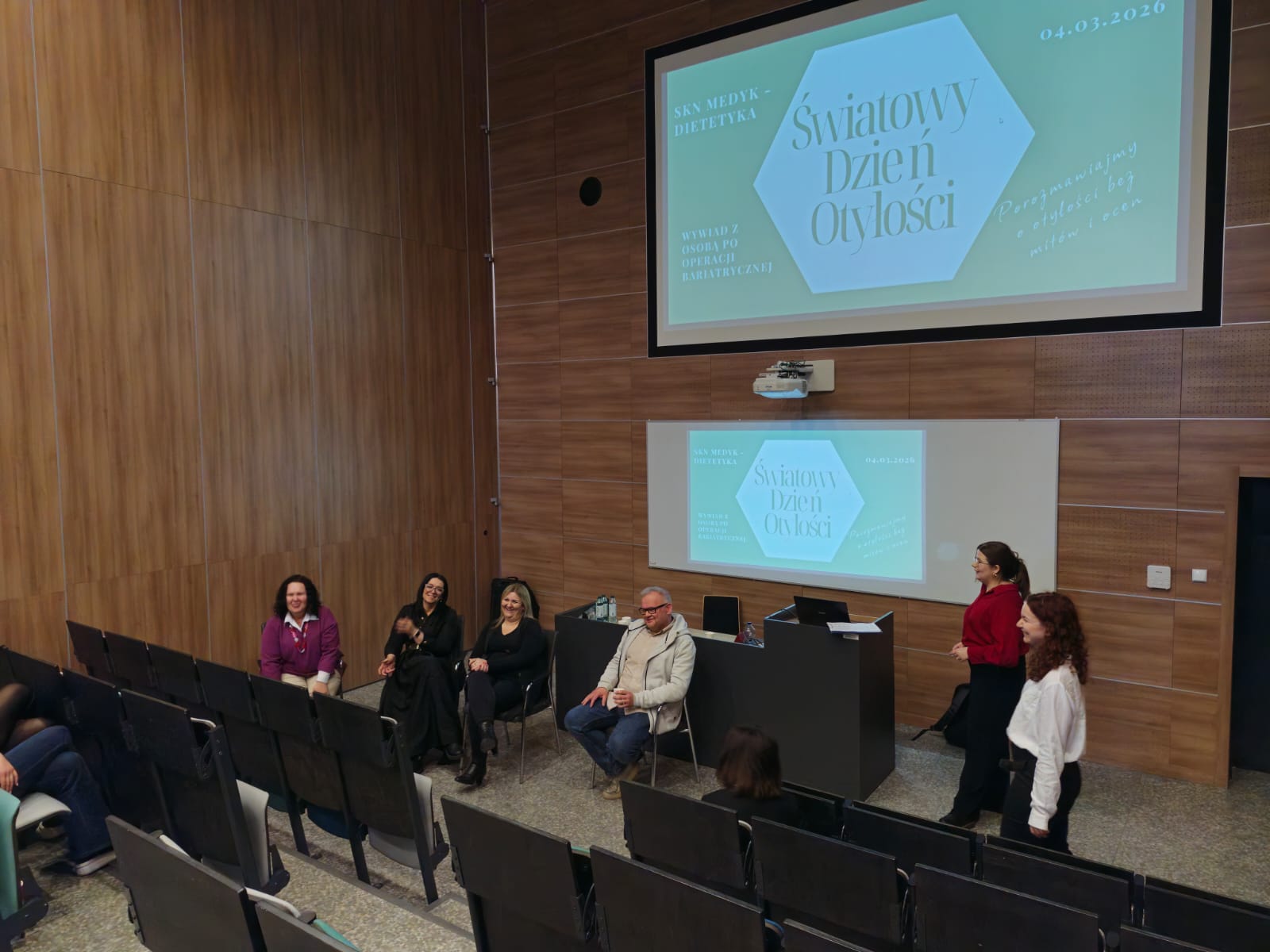 Obraz przedstawia salę wykładową z drewnianymi ścianami. Z przodu część osób siedzi na krzesłach, a jedna stoi obok mównicy. Nad nimi znajduje się duży ekran z wyświetlonym slajdem w języku polskim na jasnym tle, a poniżej mniejszy ekran powtarzający ten sam obraz. Pomieszczenie jest dobrze oświetlone.