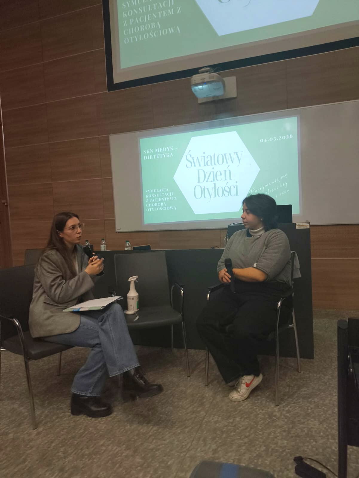 Obraz przedstawia dwie osoby siedzące i prowadzące rozmowę w sali z drewnianą ścianą. Między nimi znajduje się mały stolik z butelką. Nad nimi widoczny jest ekran z wyświetloną prezentacją oraz projektor. Pomieszczenie ma dywanową podłogę i przypomina salę wykładową lub audytorium.