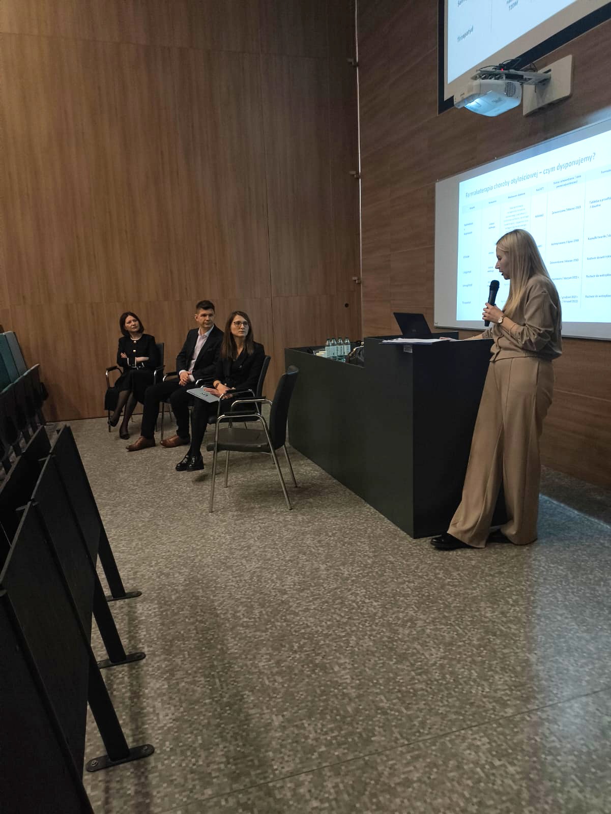 Obraz przedstawia salę konferencyjną z drewnianymi ścianami. Przy mównicy stoi osoba prowadząca wystąpienie z mikrofonem i laptopem. Z przodu siedzą uczestnicy wydarzenia, a na ekranie projektora wyświetlany jest slajd z tekstem.