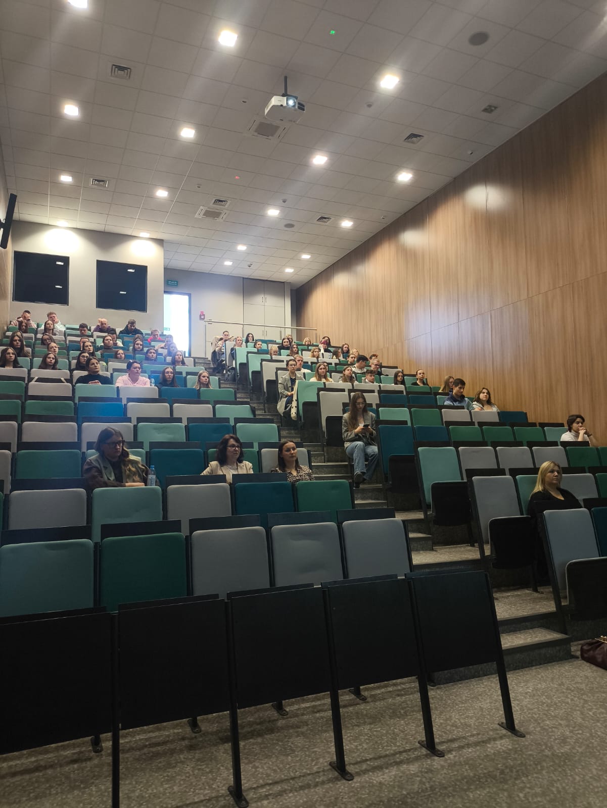 Obraz przedstawia salę wykładową z tarasowo ułożonymi miejscami, częściowo zajętymi przez uczestników. Fotele mają zielono-szare obicia, a pomieszczenie jest dobrze oświetlone sufitowymi światłami. Na suficie zamontowano projektor, a na tylnej ścianie znajdują się dwa duże ekrany. Jedna ściana jest drewniana, druga pomalowana neutralnie, a u góry widoczne jest wyjście z niebieskim znakiem.