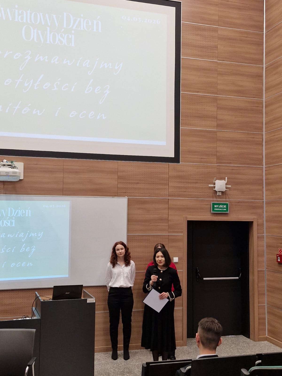 Obraz przedstawia salę wykładową z drewnianymi panelami i wyświetlonym slajdem. Z przodu, pod ekranem i tablicą, stoją osoby prowadzące prezentację, jedna z nich z mikrofonem. Publiczność siedzi naprzeciwko nich.