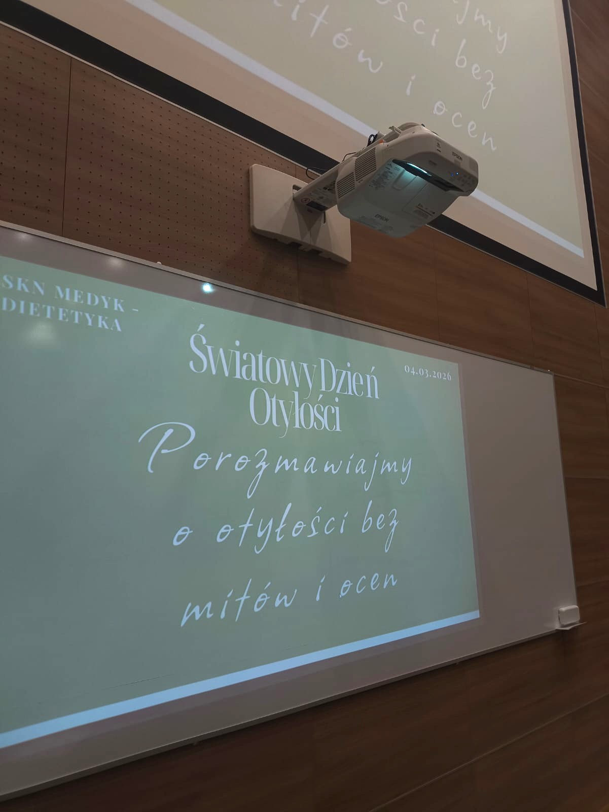 Obraz przedstawia klasę z projektorem cyfrowym wyświetlającym slajd na tablicy i ścianie. Tekst jest w języku polskim na oliwkowo-zielonym tle, a za tablicą znajduje się drewniana ściana.