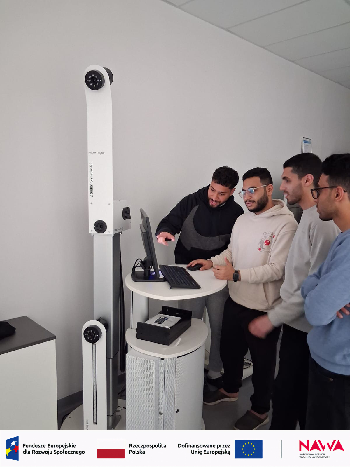 Uczestnicy stoją przy stanowisku komputerowym w nowoczesnej sali i obserwują działanie urządzenia pomiarowego z wbudowanymi czujnikami. Na stole znajduje się monitor i klawiatura, a całość wskazuje na demonstrację lub zajęcia praktyczne z wykorzystaniem specjalistycznego sprzętu.