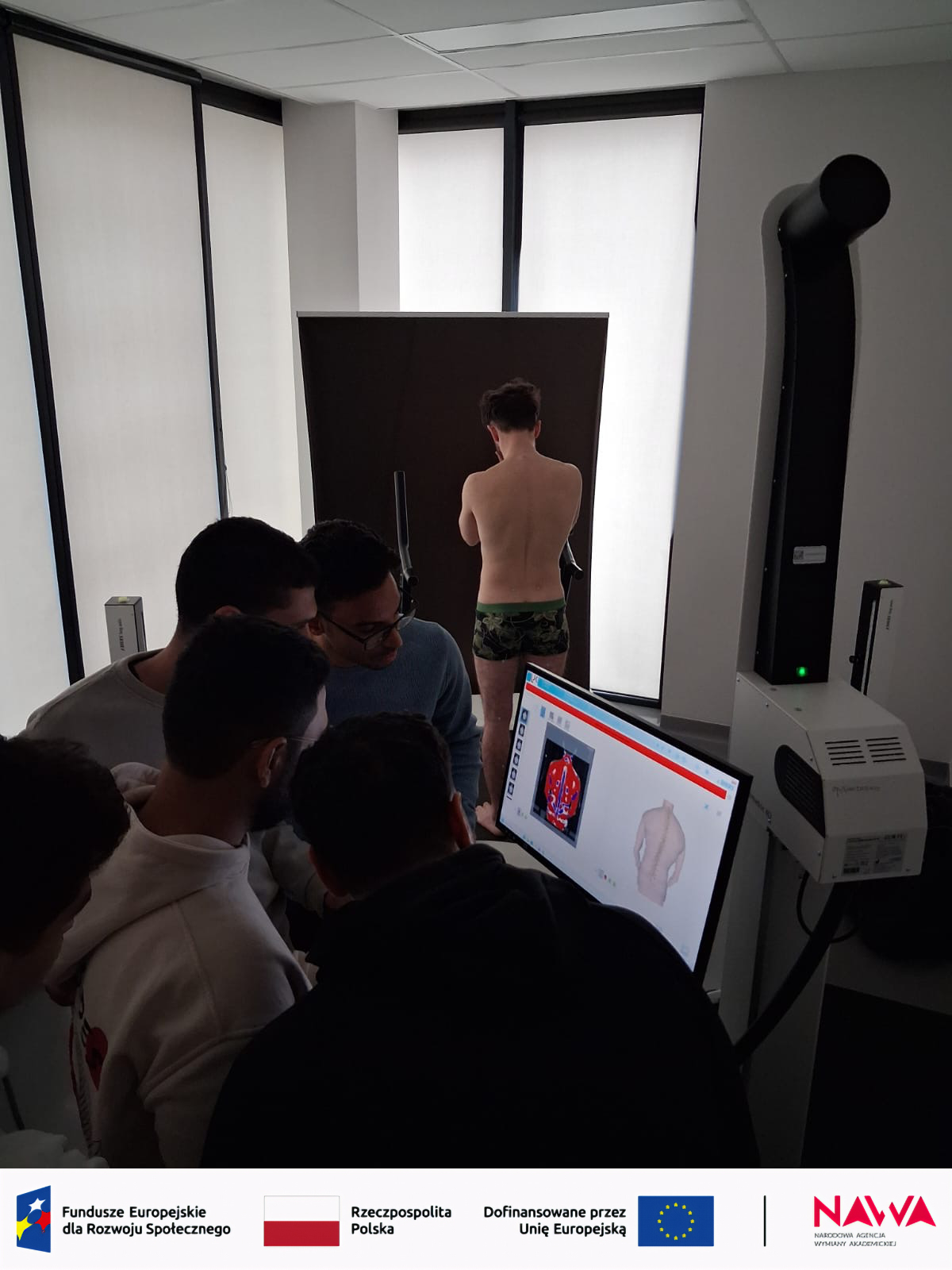 Grupa uczestników obserwuje ekran komputera w sali dydaktycznej, analizując wyniki badania prezentowane w specjalistycznym oprogramowaniu. W centrum widoczna jest osoba biorąca udział w pomiarze lub skanowaniu, a całość wskazuje na zajęcia praktyczne lub demonstrację naukową.