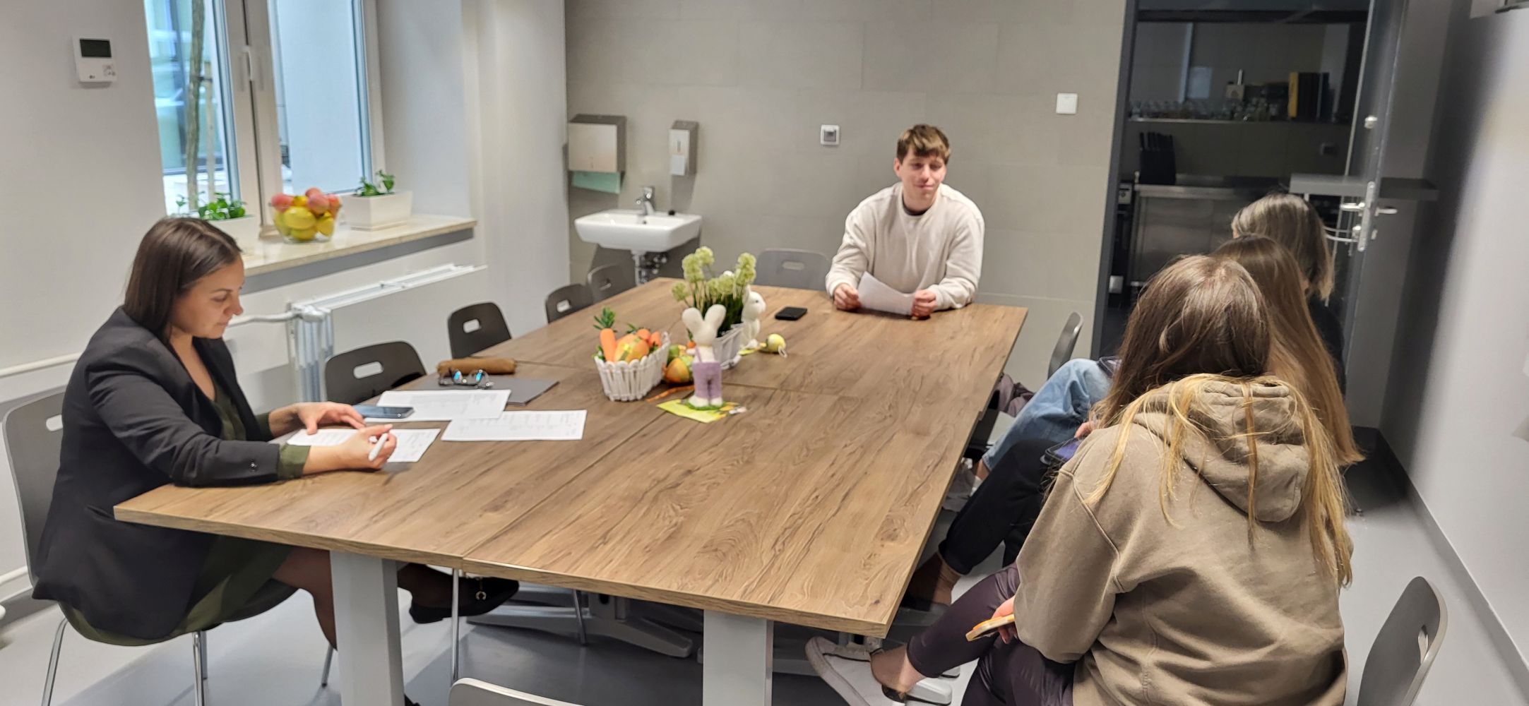 Pięć osób siedzi przy dużym stole w sali spotkań i prowadzą rozmowę lub naradę. Na stole znajdują się dokumenty, a jedna z osób sporządza notatki. Pomieszczenie jest dobrze oświetlone, z dużymi oknami, roślinami oraz elementami wyposażenia, takimi jak zlew i jasne ściany.