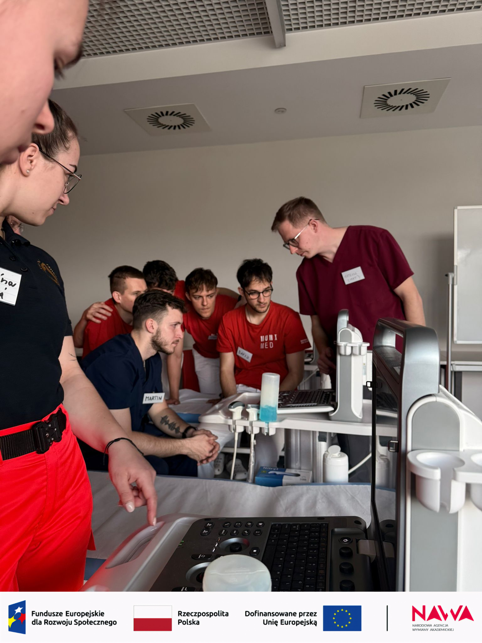Uczestnicy znajdują się w sali szkoleniowej o charakterze medycznym i obserwują demonstrację sprzętu. Jedna z osób wskazuje elementy urządzenia, omawiając jego działanie. Wokół widoczne są narzędzia i materiały, co podkreśla praktyczny charakter zajęć.