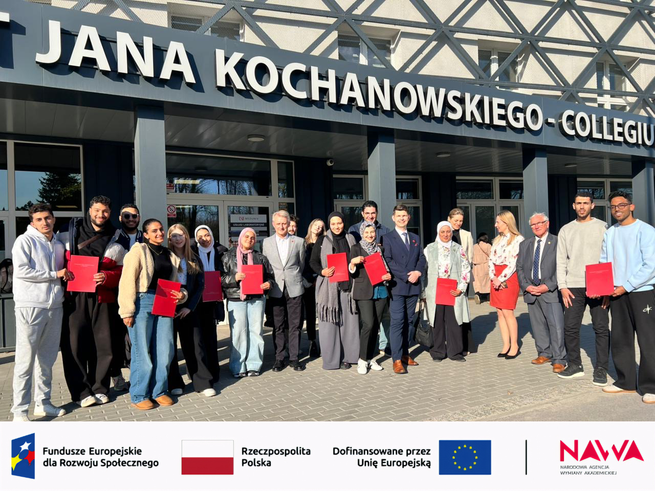 Grupa uczestników stoi przed budynkiem uczelni z widocznym napisem Collegium, pozując do wspólnego zdjęcia. Część osób trzyma materiały lub teczki, a jasne otoczenie i nowoczesna architektura wskazują na plenerowy charakter ujęcia przy obiekcie akademickim.