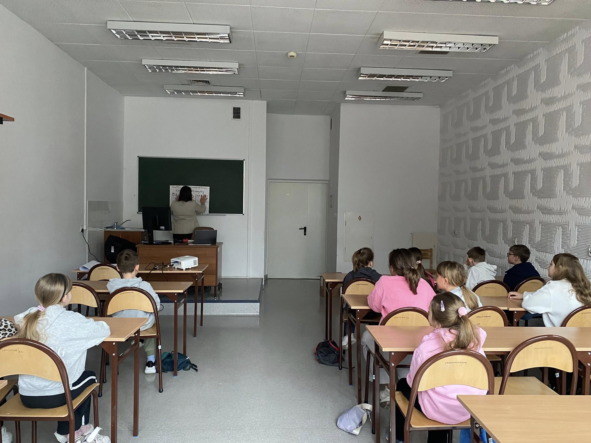 Dzieci siedzą w ławkach w sali lekcyjnej i obserwują zajęcia prowadzone z przodu. Przy tablicy znajduje się osoba prowadząca, a obok widoczne jest biurko z projektorem i sprzętem komputerowym. Pomieszczenie ma jasne ściany, oświetlenie sufitowe oraz podstawowe wyposażenie szkolne.