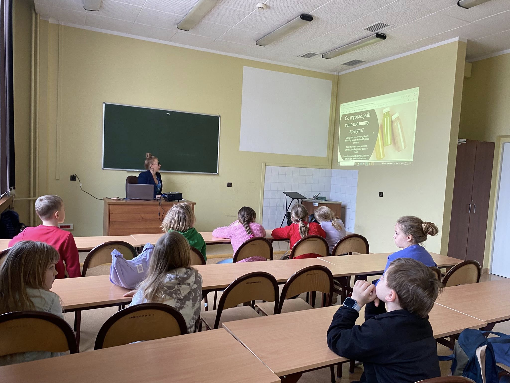 Sala lekcyjna, w której dzieci siedzą przy ławkach i obserwują prezentację wyświetlaną na ekranie. Osoba prowadząca pracuje przy biurku z laptopem, kierując zajęciami. Wnętrze ma jasne ściany, tablicę, szafkę oraz oświetlenie sufitowe, co wskazuje na standardowe środowisko szkolne.